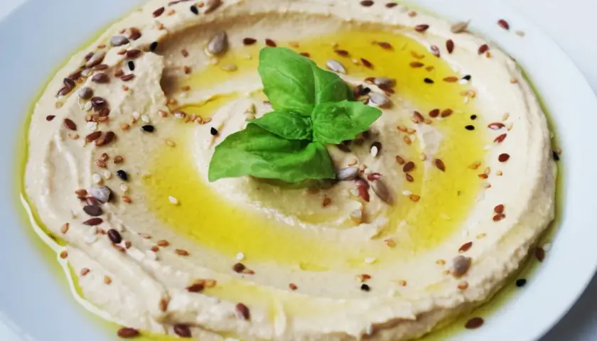 Hummus di ceci cremoso, condito con olio d'oliva, semi misti e una foglia di basilico. Perfetto da gustare con pane pita o verdure fresche.