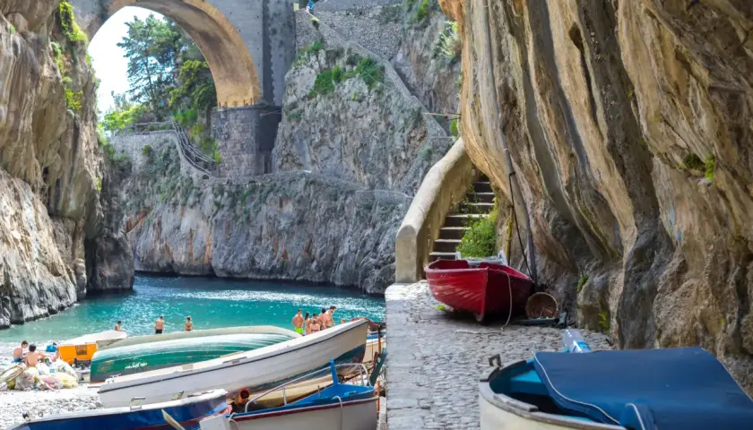 Boats line the shore of Fiordo di Furore, a stunning fjord with turquoise waters and a dramatic arched bridge overhead.