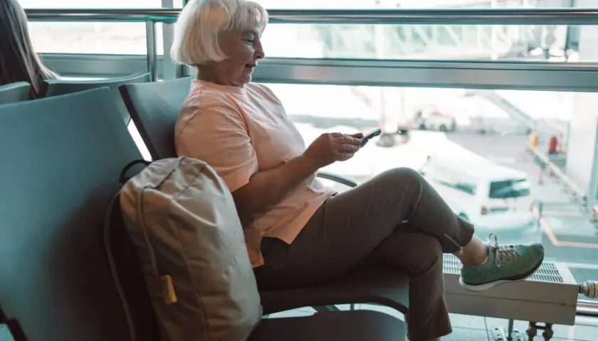 Uśmiechnięta starsza pani z siwymi włosami, korzystająca z telefonu w poczekalni lotniska. Jej plecak leży obok. To komfortowy lot samolotem osoby niepełnosprawnej.