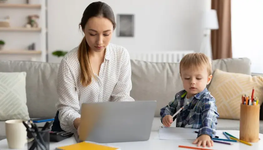 Kobieta pracuje na laptopie, podczas gdy jej syn rysuje obok. To codzienna opieka na dziecko i praca zdalna w jednym.