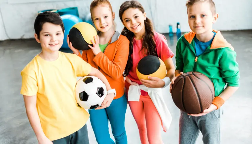 Cztery uśmiechnięte dzieci w sportowych ubraniach trzymają piłki: nożną, do koszykówki i dwie treningowe.