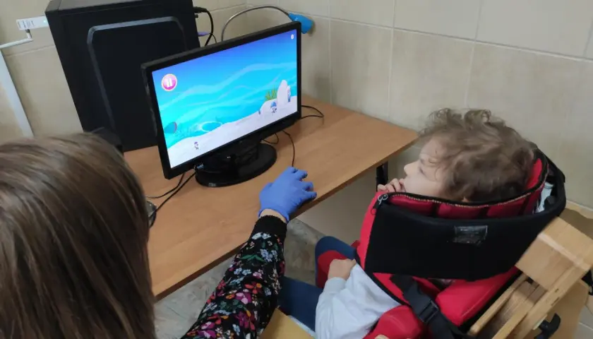 Dziecko w specjalnym foteliku siedzi przed komputerem, na ekranie gra. Obok siedzi osoba w rękawiczce.