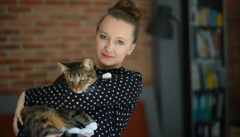 Kobieta w czarnej bluzce w kropki trzyma na rękach pręgowanego kota. To jej profilowe zdjęcie z kotem.