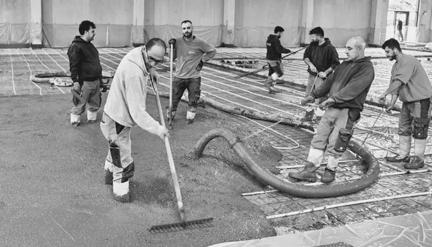 Arbeiter verteilen frisch gegossenen stahlfaserbeton bodenplatte mit Rechen. Beton wird aus Schlauch gepumpt.