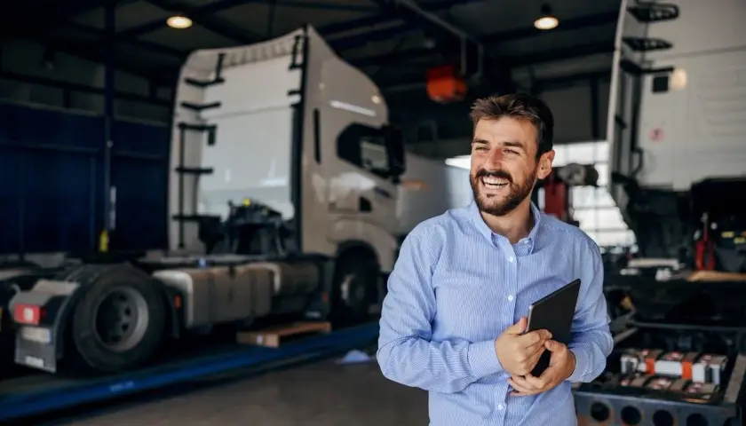 Uśmiechnięty mężczyzna z tabletem w ręku, w tle warsztat z ciężarówkami. Oferuje **usługę transportową ryczałt**.