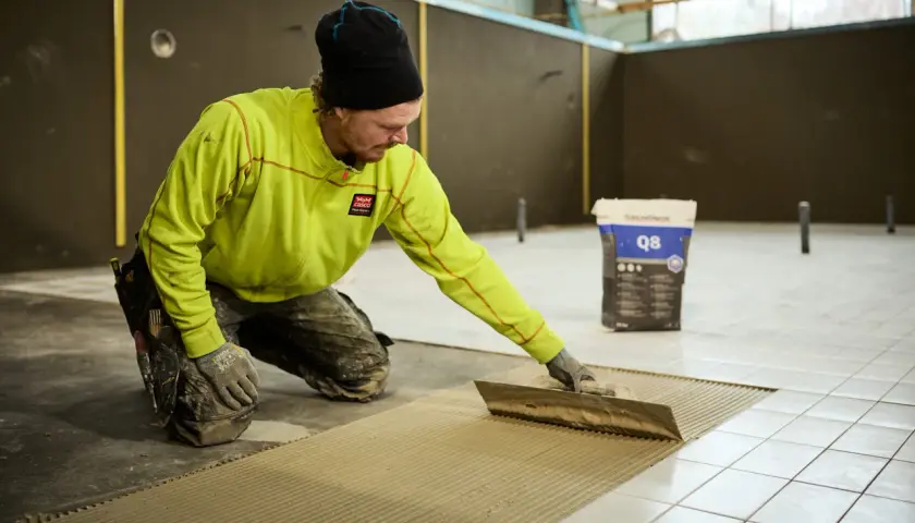 Man i gul jacka och mössa sprider ut fix med en tandspackel för att byta golv. En säck med fix står i bakgrunden.