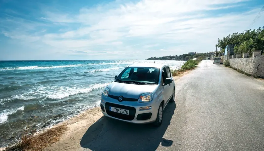 A silver Fiat Panda parked on a coastal road in Greece. The turquoise sea laps the shore beside the car, suggesting this is the perfect way to explore.