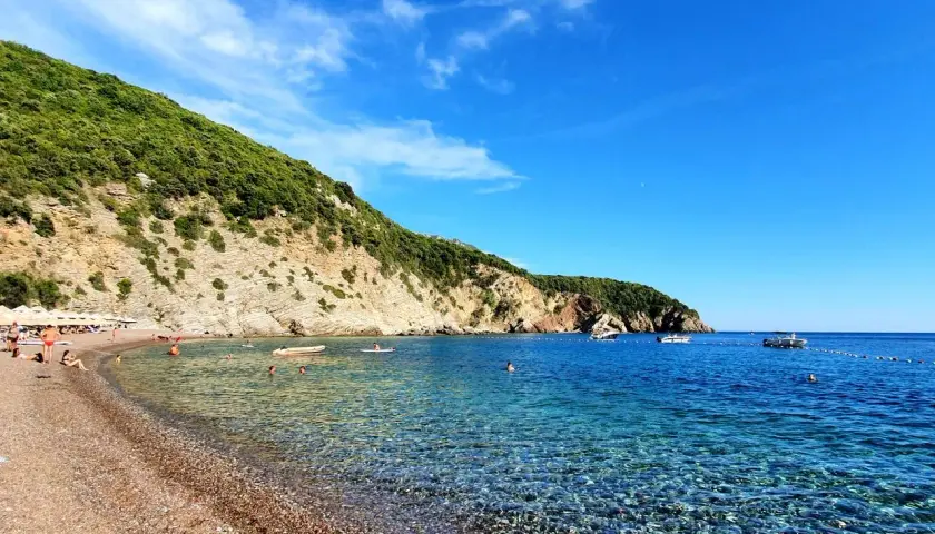 Kamienista plaża w Czarnogórze z turkusową wodą i zielonym wzgórzem w tle.