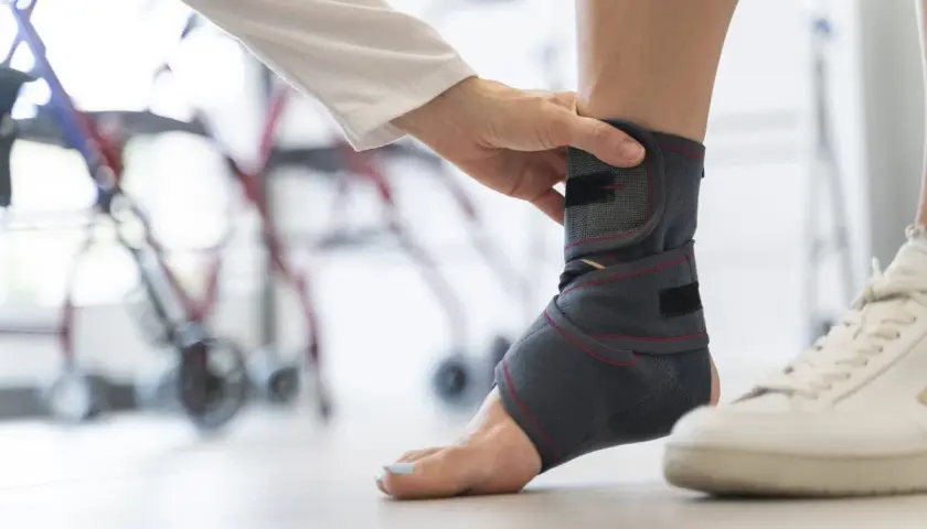 Dłoń terapeuty dopasowuje stabilizator stawu skokowego. W tle widać rehabilitacyjny sprzęt.