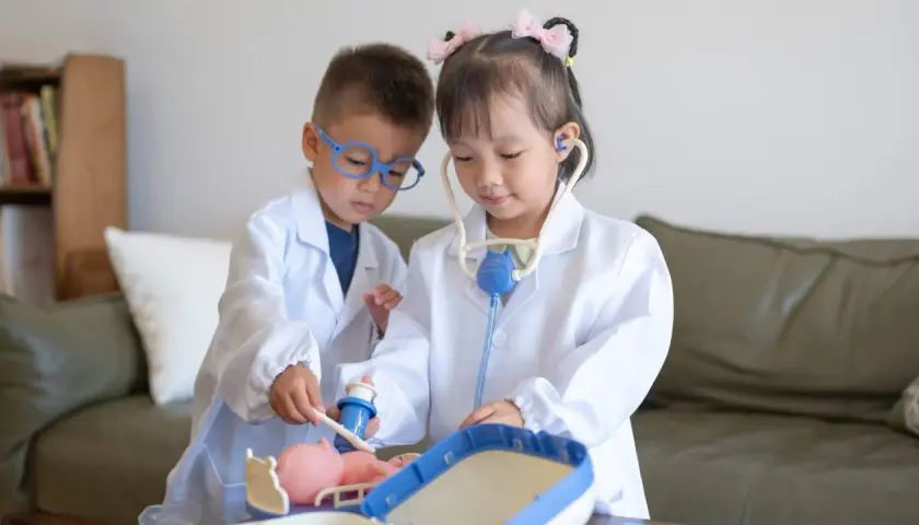 Dos niños, vestidos de doctores, atienden a un muñeco. La niña usa un estetoscopio, mientras el niño le administra una inyección. Es una escena de juego de roles, una brecha de Rolando con niños.