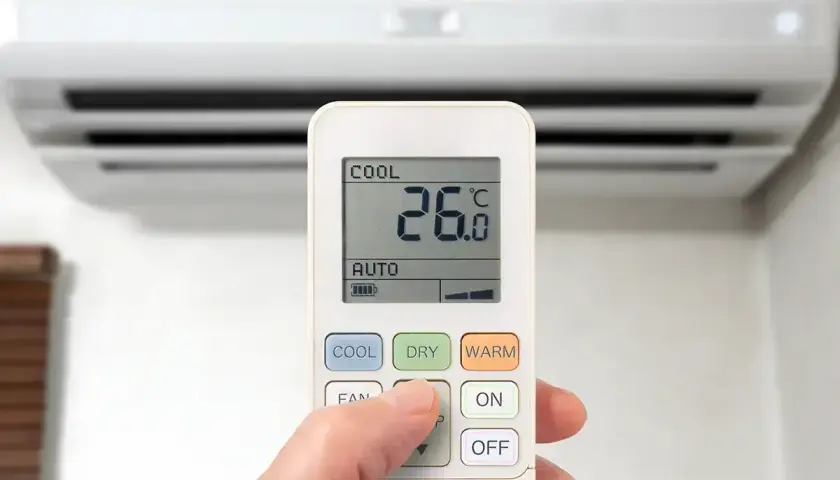 Dłoń naciska przycisk na pilocie klimatyzacji, wybierając funkcje chłodzenia. Na ekranie widoczna temperatura 26°C.