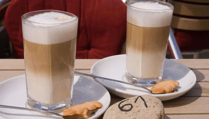Dwa latte macchiato, czyli kawy z warstwami mleka i pianki, na stoliku. Obok ciasteczka i kamień z numerem 61.