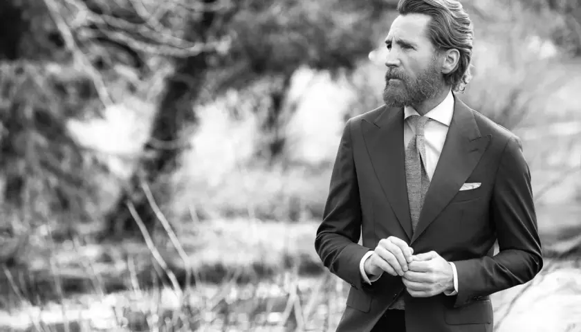 Un homme barbu en costume sombre, chemise blanche et cravate grise, ajuste sa veste. Sa tenue avec short blanc homme est élégante.