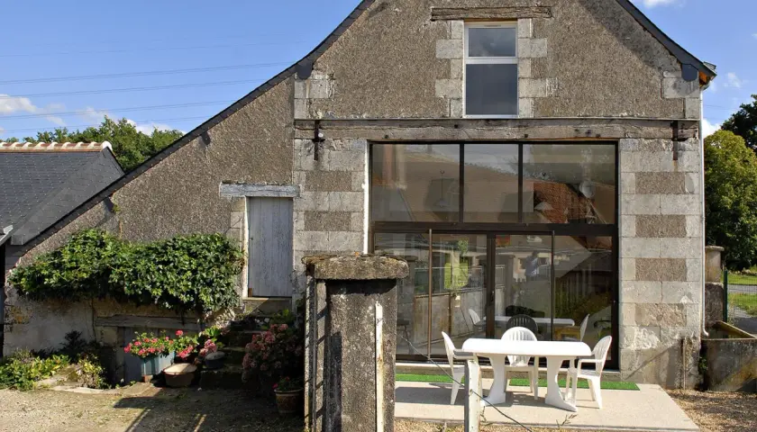Ancienne grange en cours de rénovation, avec une façade en pierre et un grand mur vitré moderne. Une table et des chaises blanches sont installées sur la terrasse.