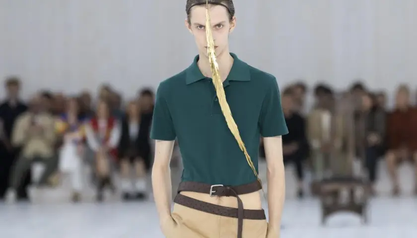 Enzo Vogrincic, modelo con un polo verde, pantalones caqui y un adorno dorado en la cara, desfila ante un público difuminado.