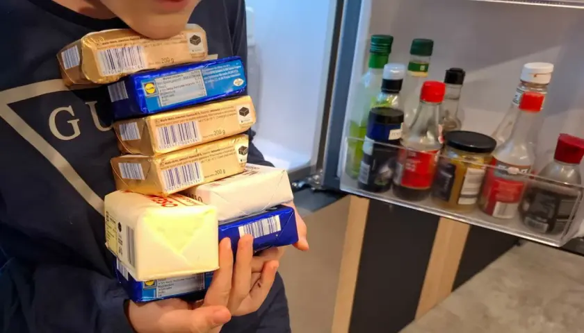 Dziecko trzyma stos opakowań masła przed otwartą lodówką z butelkami sosów.