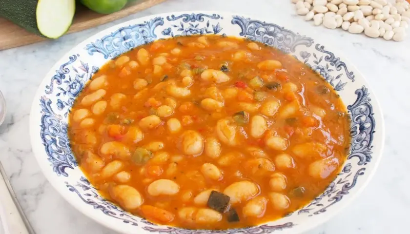 Un plato de alubias con verduras, cocinadas en una salsa rojiza y servidas en una vajilla con borde azul.