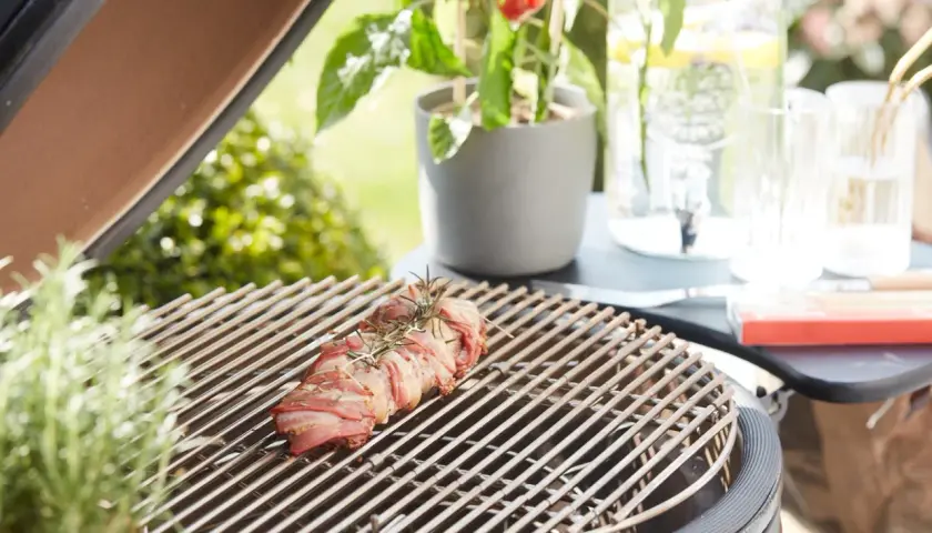 Un rôti de viande enroulé de lard, prêt à griller. Une légère **fume bleu** s'échappe, promettant un repas savoureux en plein air.