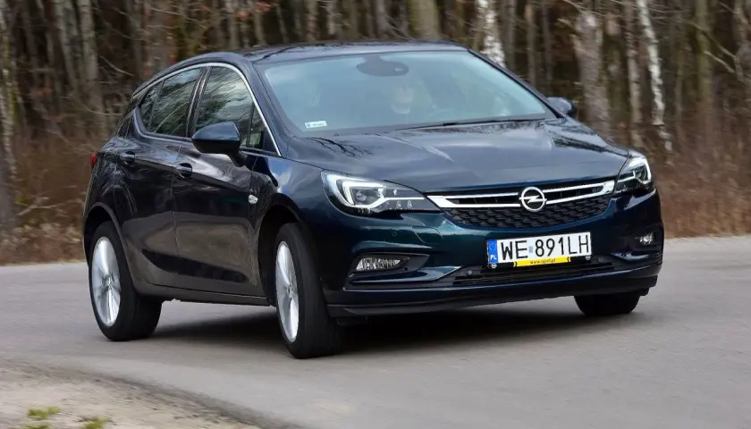 Ciemnozielony Opel Astra K 1.4 Turbo jedzie po drodze wśród drzew.