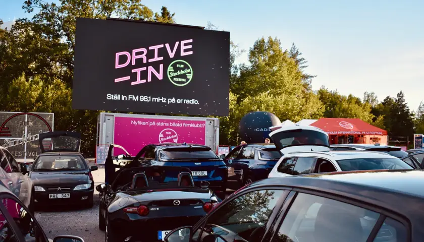 Bilar samlade vid en solvalla drive in bio. Stor skärm visar "DRIVE-IN FILM Stockholm Festival".