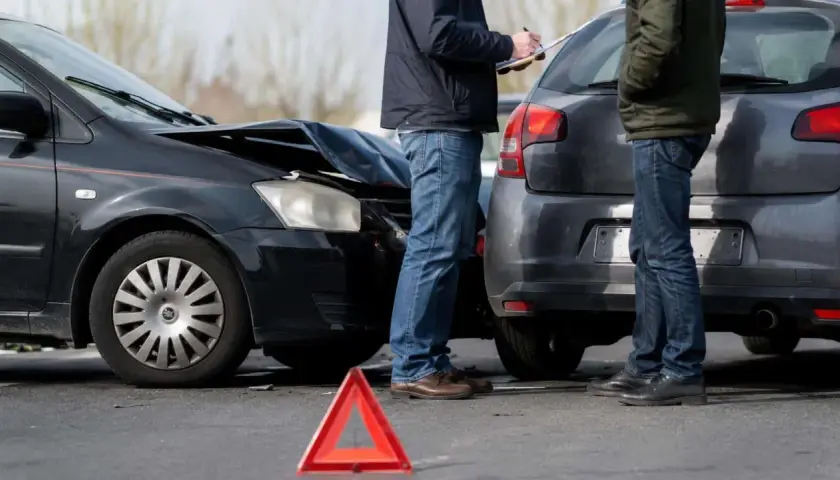 Dwaj mężczyźni spisują protokół po kolizji. Zniszczona maska czarnego auta i czerwony trójkąt ostrzegawczy sugerują, że czeka ich mandat za kolizje.