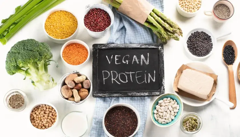 Tablica z napisem "VEGAN PROTEIN" otoczona różnorodnymi roślinnymi źródłami białka: soczewicą, fasolą, brokułem, szparagami, tofu i orzechami.