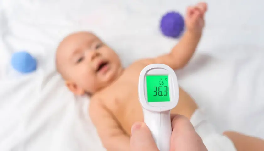 Dziecko leży na białym tle, a ręka trzyma termometr bezdotykowy. Na ekranie termometru widoczna jest temperatura 36.3°C, co oznacza, że temperatura mierzona w odbycie jest prawidłowa.