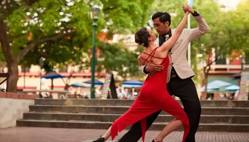 Para tańczy tango, jej czerwona sukienka faluje, a on w jasnej marynarce. W tle drzewa i schody.