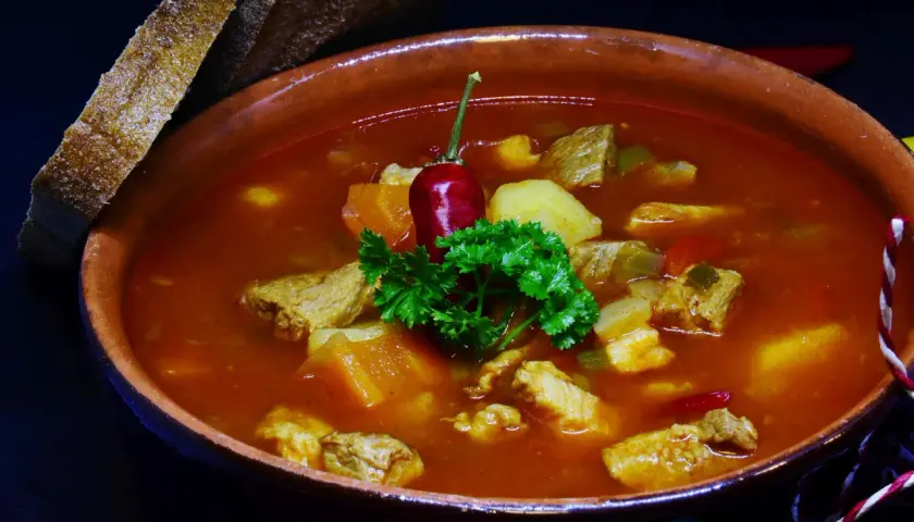 Gęsty bogracz wieprzowy z kawałkami mięsa, ziemniaków i papryki, ozdobiony papryczką chili i natką pietruszki. Obok kromki chleba.