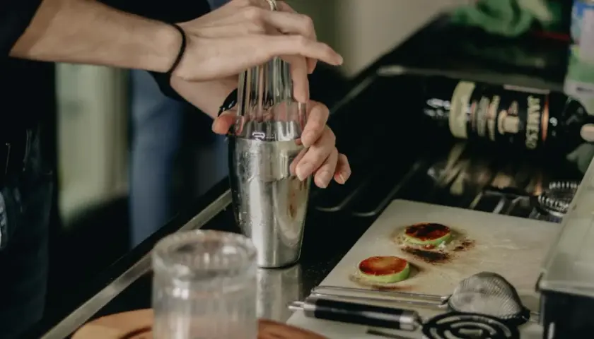 Ręce mieszają składniki w shakerze, przygotowując drinki na ciepło. Na blacie leży butelka whiskey i przypalone plasterki jabłka.