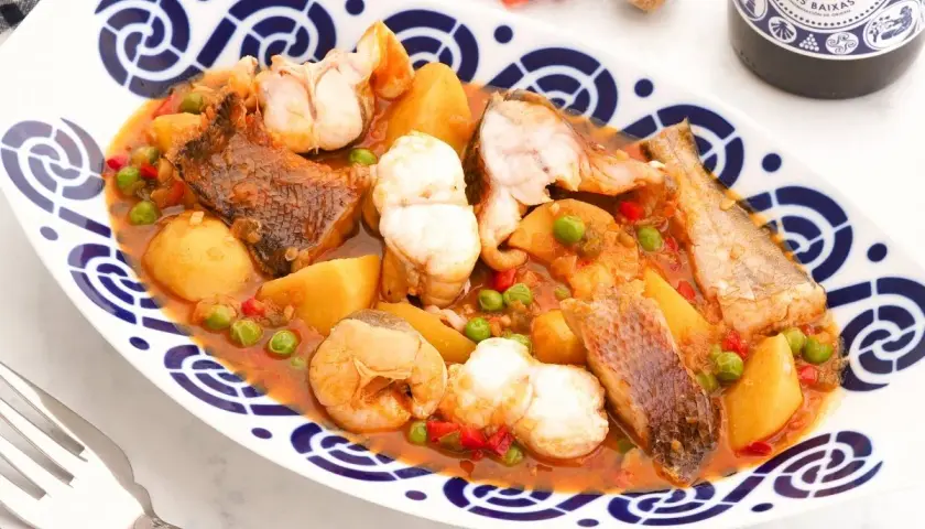 Caldeirada gallega servida en plato blanco con borde azul. Contiene trozos de pescado, patatas, guisantes y pimientos en una salsa rojiza.