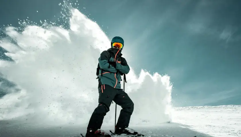 Narciarz w zielono-czarnej kurtce snowboardowej, z pomarańczowymi akcentami, w kasku i goglach, stoi na nartach, a za nim wybucha śnieżna lawina.