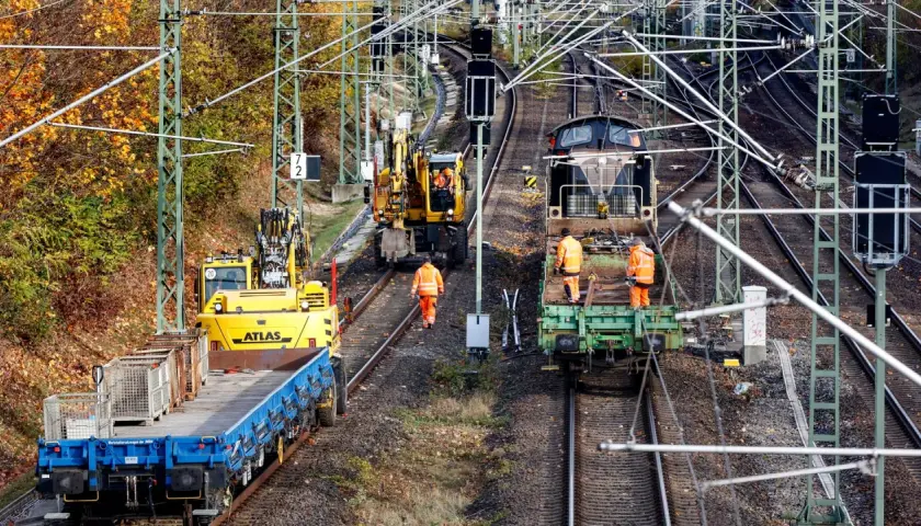 Arbeiter in orangefarbenen Warnwesten inspizieren Gleise neben Baumaschinen und Zügen. Die Szene zeigt die Infrastruktur der serbischen Eisenbahnen im Herbst.