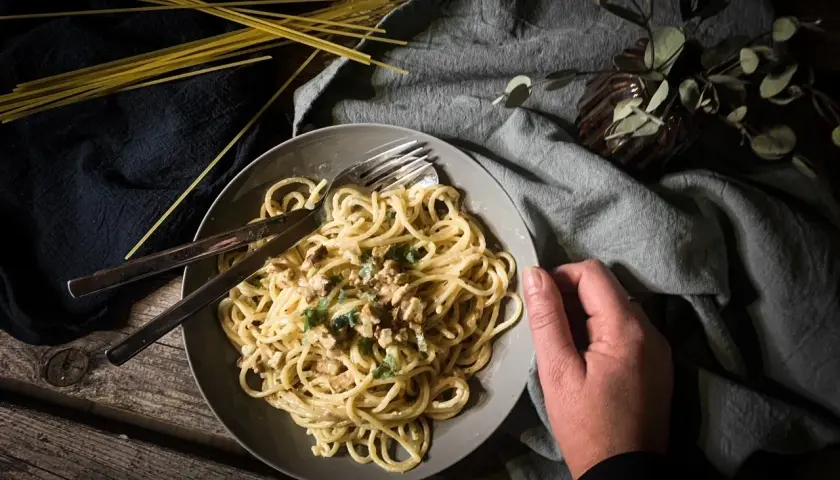 Teller mit cremiger, veganer Spaghetti Carbonara, garniert mit Kräutern. Daneben liegen ungekochte Spaghetti.