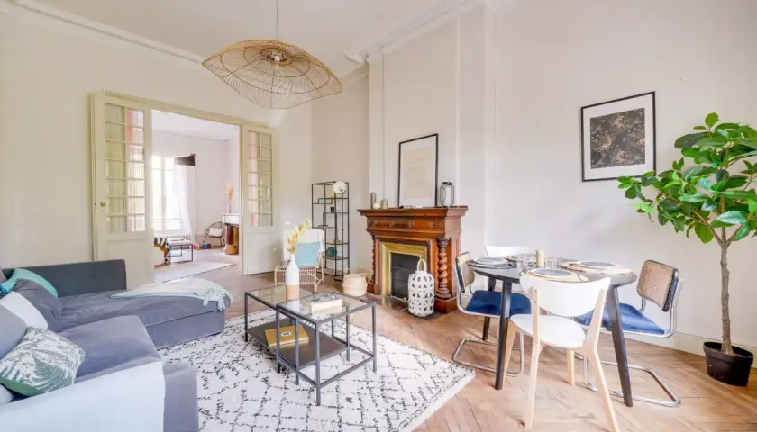 Salon lumineux avec canapé gris, tapis graphique, cheminée ancienne et table à manger. Une belle idée home staging pour un espace convivial.