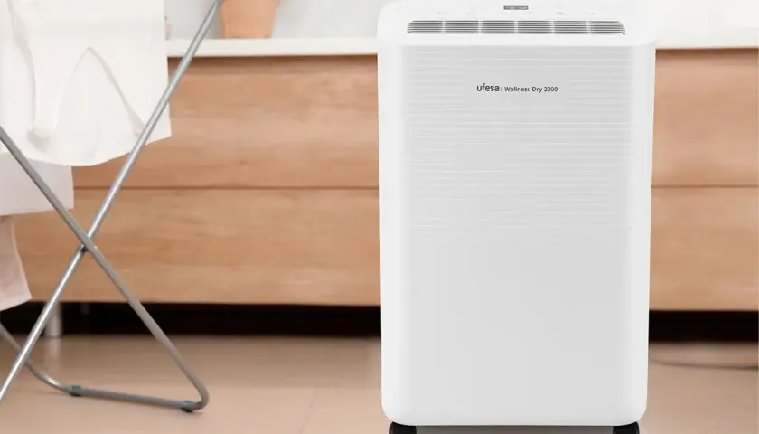 Un deshumidificador Ufesa Wellness Dry 2000, blanco y con ruedas, junto a un tendedero con ropa.