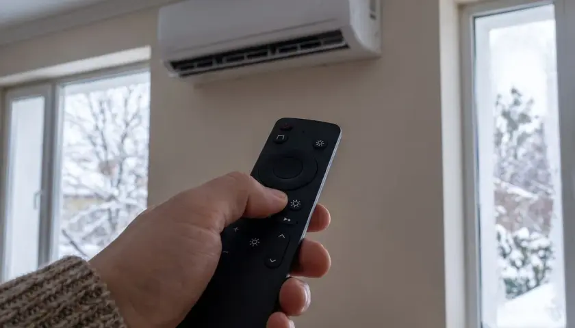 Mano con mando a distancia ajustando el icono de calor en aire acondicionado. Afuera, ventanas con nieve.
