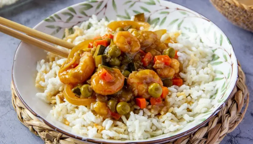 Un plato de arroz blanco cubierto con gambas en salsa agridulce y verduras. Los palillos descansan sobre el borde del cuenco.