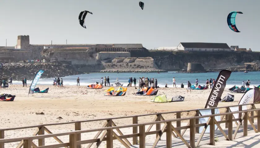 Plaża pełna kolorowych latawców i ludzi, gotowych do **kitesurfingu w Polsce**. W tle widać zabytkowe budowle.