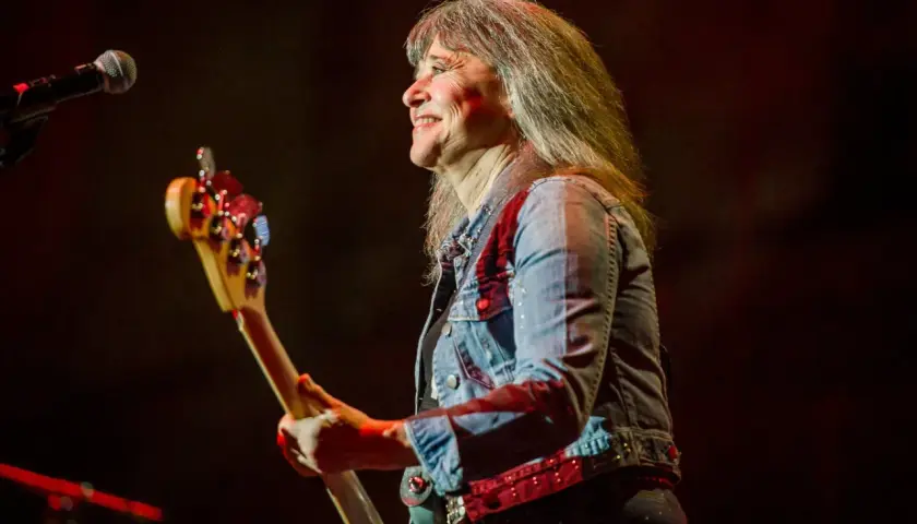 Suzi Quatro obecnie, z uśmiechem na twarzy, gra na gitarze basowej w dżinsowej kurtce.
