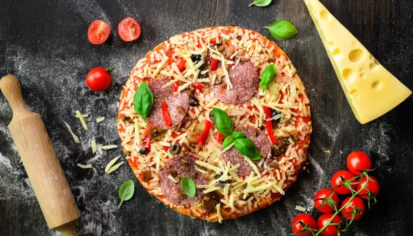 Gotowa pizza z salami, serem i warzywami, obok wałek, ser i pomidorki. Idealna do przygotowania z odpowiednią mąką do pizzy.