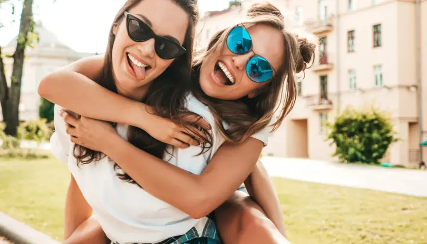 Dos amigas riendo y abrazándose, como si estuvieran escribiendo una carta a una amiga. Llevan gafas de sol y están al aire libre.