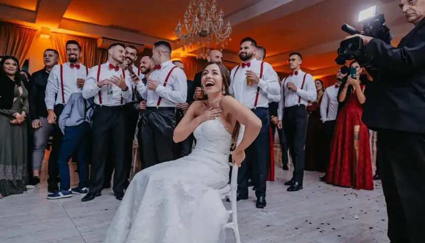 Radosna panna młoda na krześle, otoczona przez drużbów w czerwonych szelkach. Zastanawiasz się, ile kosztuje wesele na 50 osób?