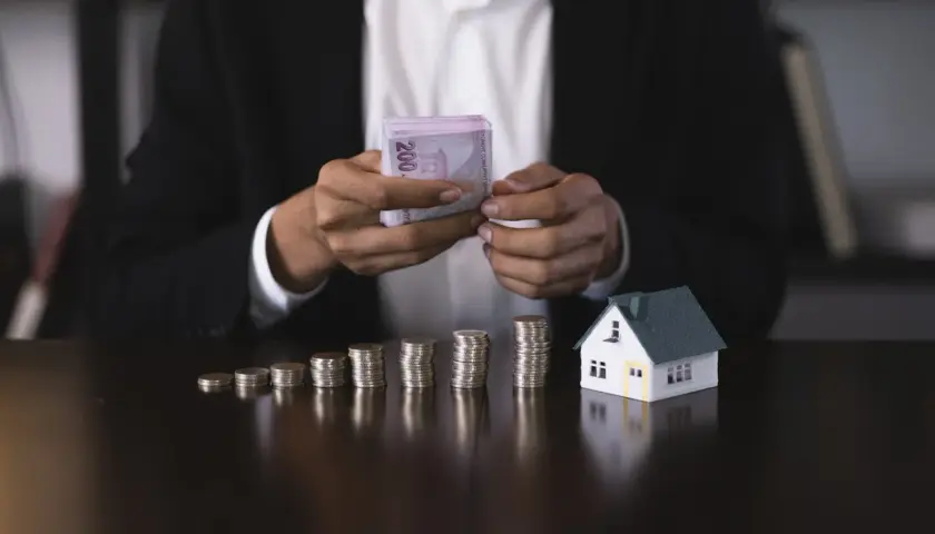 Ręce w garniturze liczą pieniądze obok modelu domu i stosów monet. Czy to zadatek, czy zaliczka na wymarzone mieszkanie?