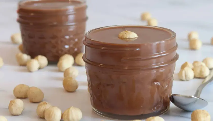 Dos tarros de deliciosa nutella vegana rodeados de avellanas. Una cucharilla plateada descansa cerca.