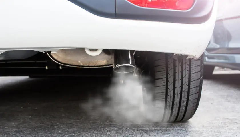 Białe auto pluje wodą z wydechu, tworząc mgiełkę na asfalcie. Widać fragment opony i tylną lampę.