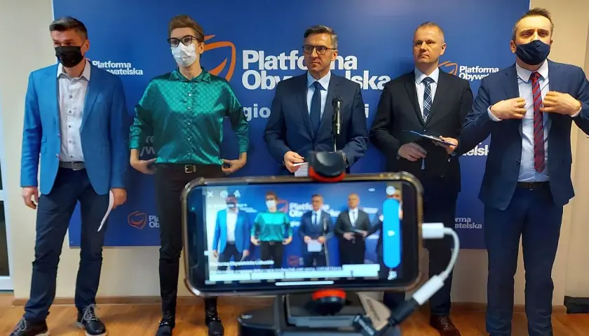 Pięciu mężczyzn w garniturach stoi przed niebieskim banerem z logo Platformy Obywatelskiej. Na pierwszym planie widać telefon nagrywający wydarzenie.