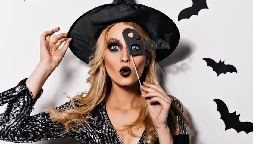 Kobieta w kapeluszu czarownicy z literami "Boo!" przed twarzą. Jej prosty makijaż czarownicy podkreśla oczy i usta.