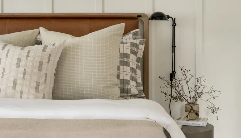 Studio McGee bed styling with layered pillows featuring subtle patterns and a plaid accent. A bedside lamp and delicate branches in a vase complete the serene look.