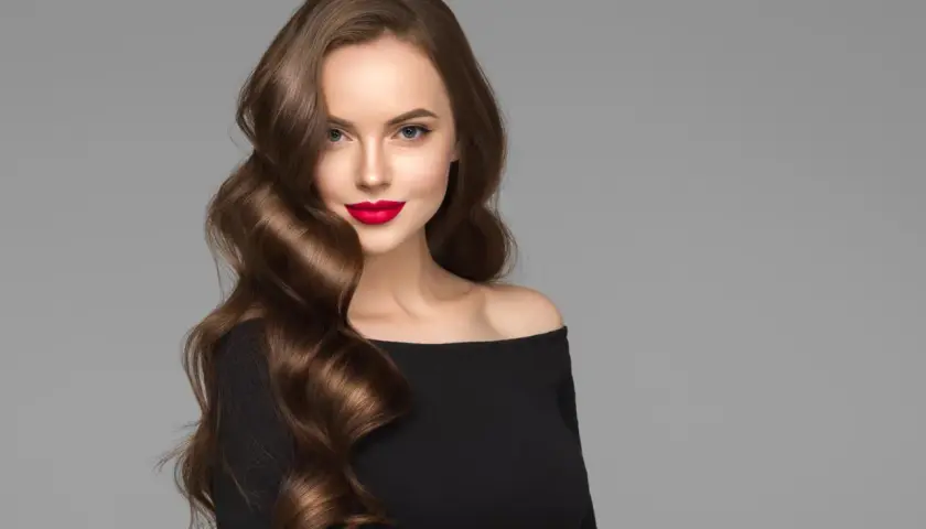 Mujer con labios rojos y espectaculares ondas pelo largo castaño, luciendo un vestido negro de hombros descubiertos.